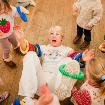 A playful group of young children surrounds a woman lying on the wooden floor, laughing and engaging in an interactive activity. The woman, wearing a white outfit, has colorful textured rings on her arms and legs, while the children, dressed in warm clothing, excitedly participate. The setting appears to be an indoor play or learning environment.